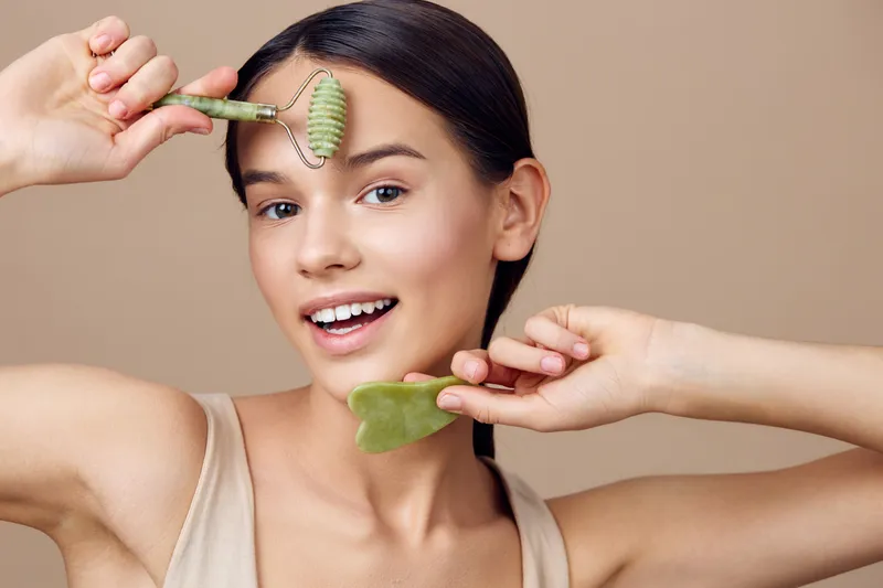 Young woman with facial roller showcasing skincare routine, smiling and holding a gua sha tool, on a neutral background, promoting beauty and self care