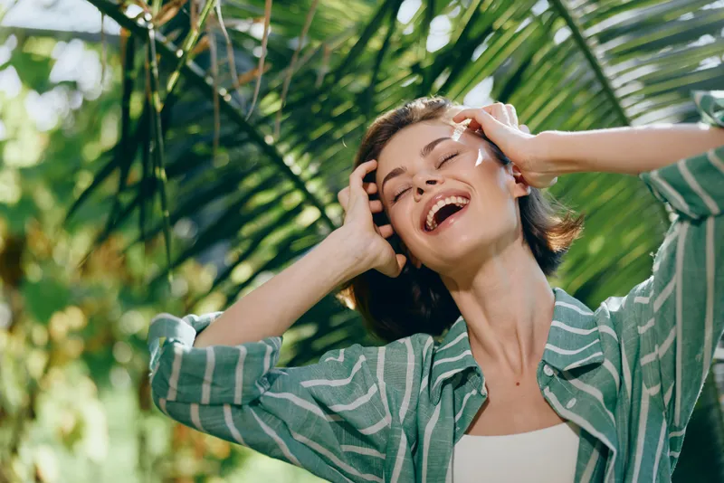 Joyful woman outdoors, smiling with hands in hair, under bright sunlight amid lush greenery and tropical leaves.