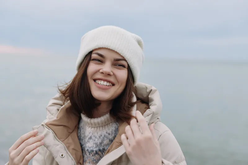Woman portrait outdoors in winter, smiling in a warm jacket and coat, beanie, beside a calm sea, capturing casual fashion and relaxed mood.