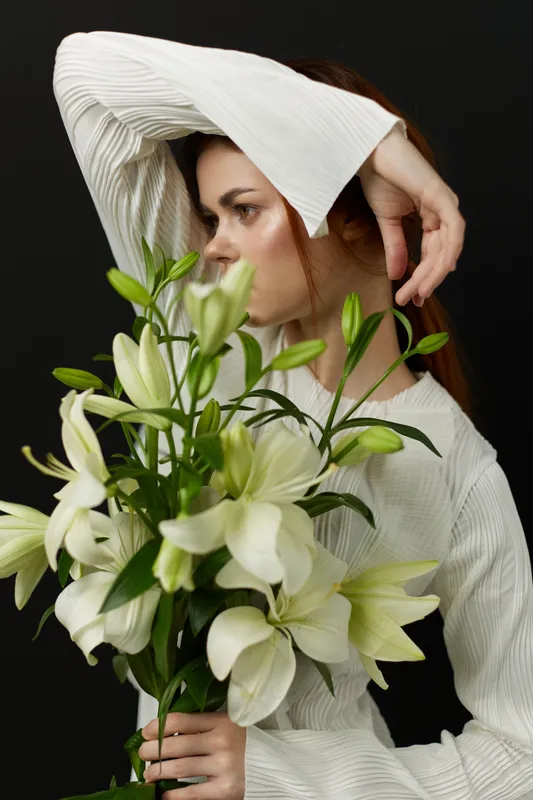 Young woman in a white outfit holding white lilies against a dark background, expressing serenity and elegance in a floral composition