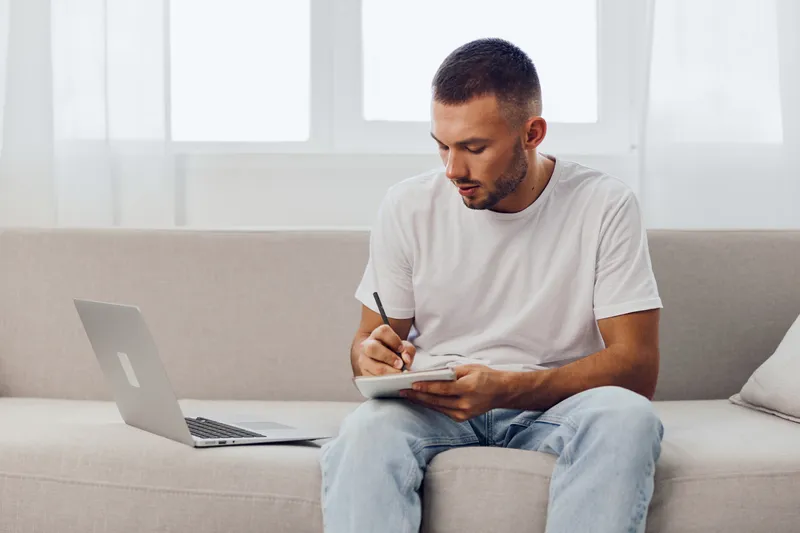 Young man working on a laptop while taking notes, creating a productive atmosphere in a modern home office setting, bright and minimalistic design.