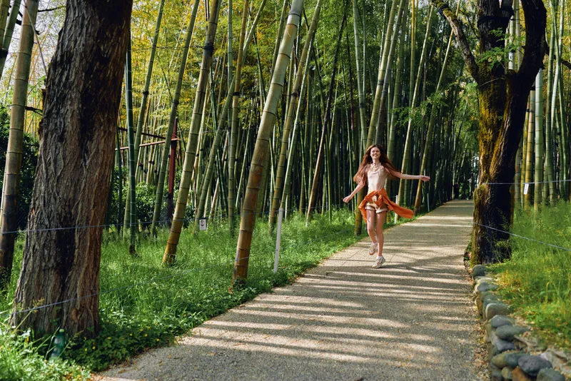 Woman running on a gravel path through a dense bamboo forest, wearing a flowing dress with arms outstretched, joyful expression and sunlight filtering through tall trunks and green foliage.