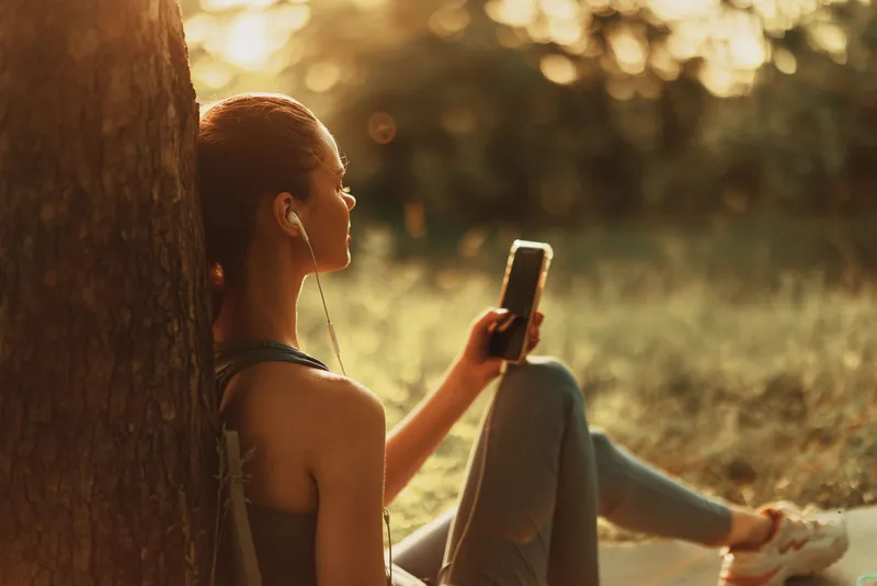 Young woman enjoying music outdoors in a summer evening, sitting against a tree with warm sunlight providing a serene atmosphere, showcasing relaxation and tranquility