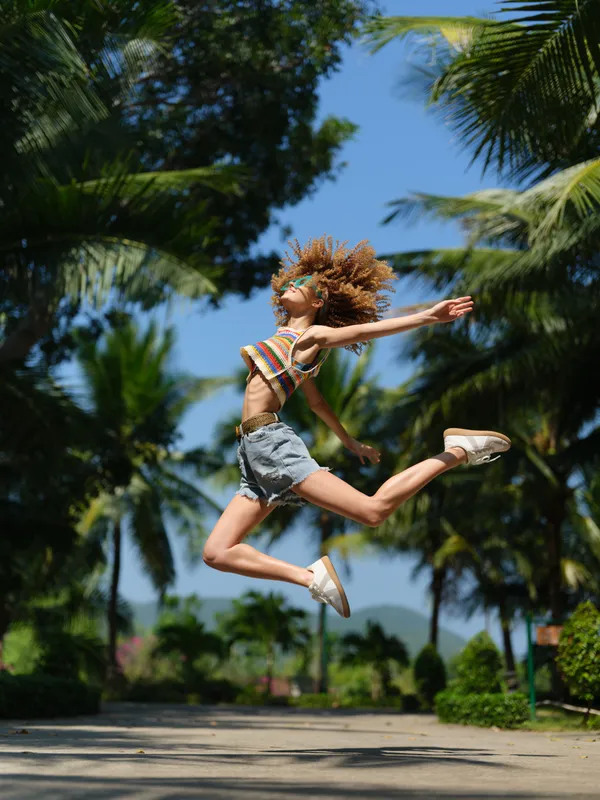 joyful woman jumping outdoors in summer, wearing colorful top and denim shorts with curly hair, surrounded by palm trees under clear blue sky.