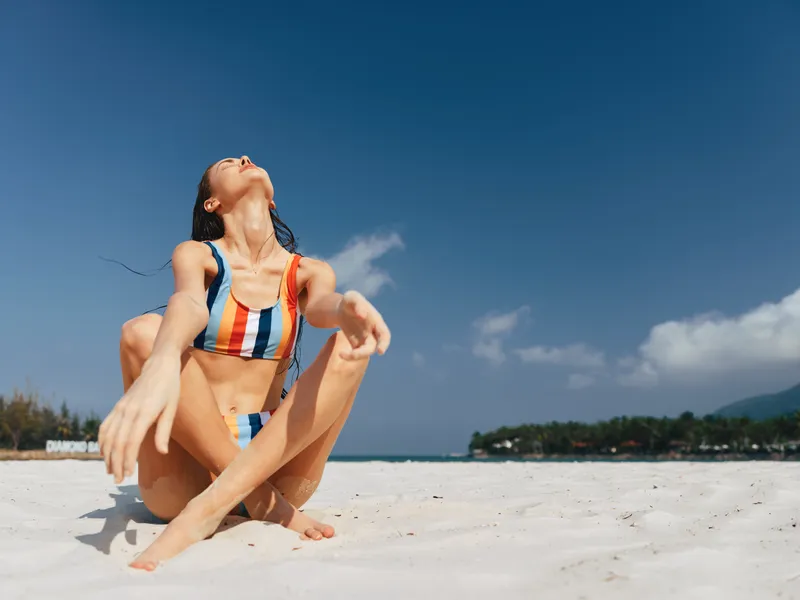 Sunlit beach scene shows a graceful woman in a striped bikini sitting on white sand, arms extended, head tilted back, basking in warm rays and crystal blue sea.