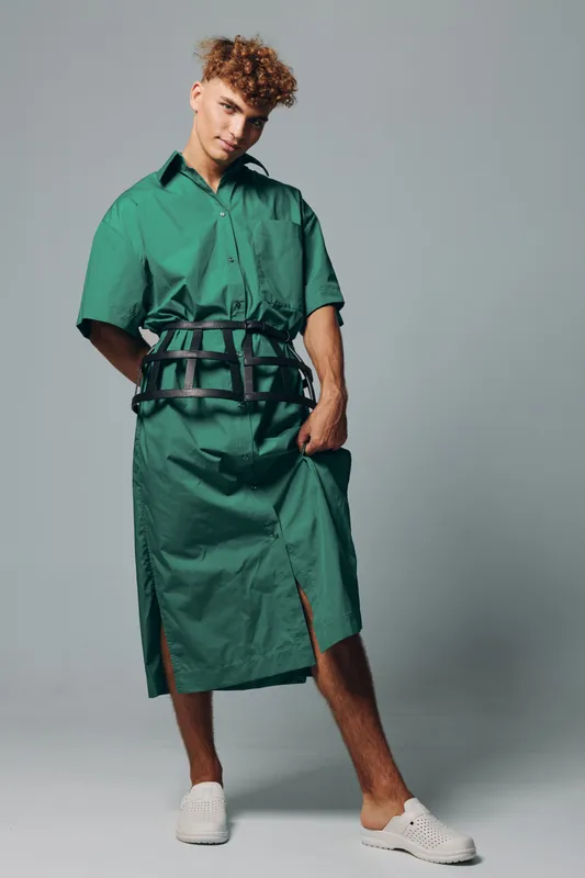 Young man posing in green oversized shirt dress with black harness belt and white sneakers against plain background. Fashion model with curly hair and casual confident stance.