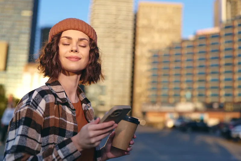 Woman with coffee and phone in city during golden hour, smile and candid authenticity in a mindful lifestyle moment, golden hour glow and emotional storytelling in portrait.