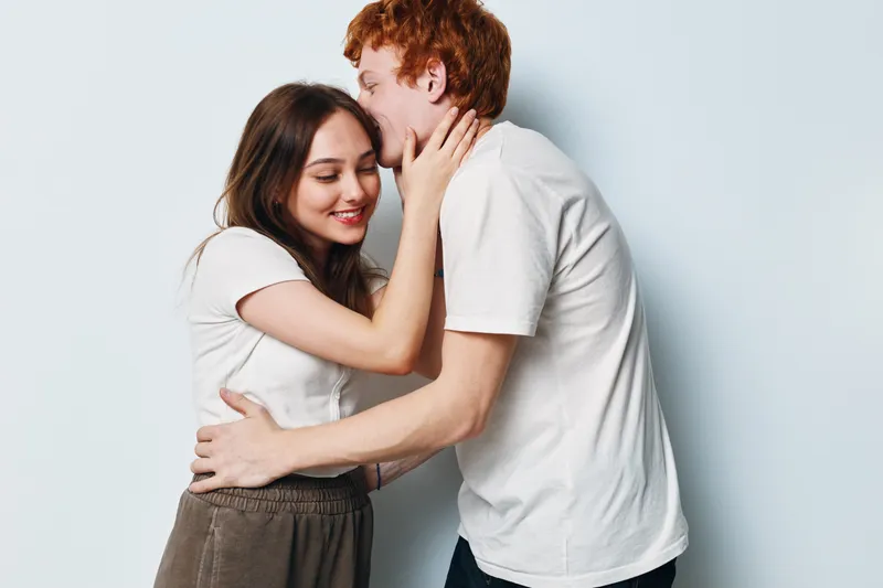 Couple, love, embrace, and affection with man kissing woman on cheek in studio. Happy relationship, casual clothing, and smiling young people for romance and intimacy.
