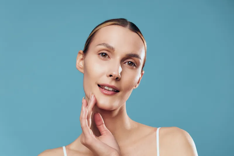 Young woman skincare portrait, close up face, smooth skin, touching cheek with fingertips, beauty and wellness concept, spa mood, soft light, isolated on blue background, copy space