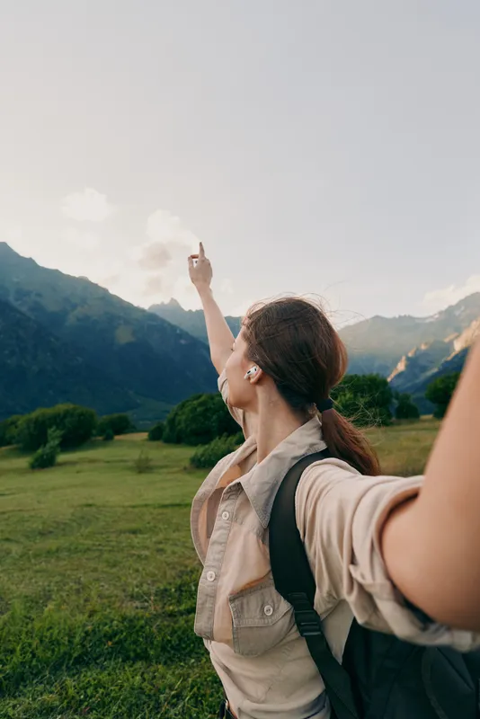 Smiling woman with backpack posing outdoors in a mountainous landscape, pointing toward the sky, capturing a lively travel moment filled with exploration and wellness and a sense of freedom.