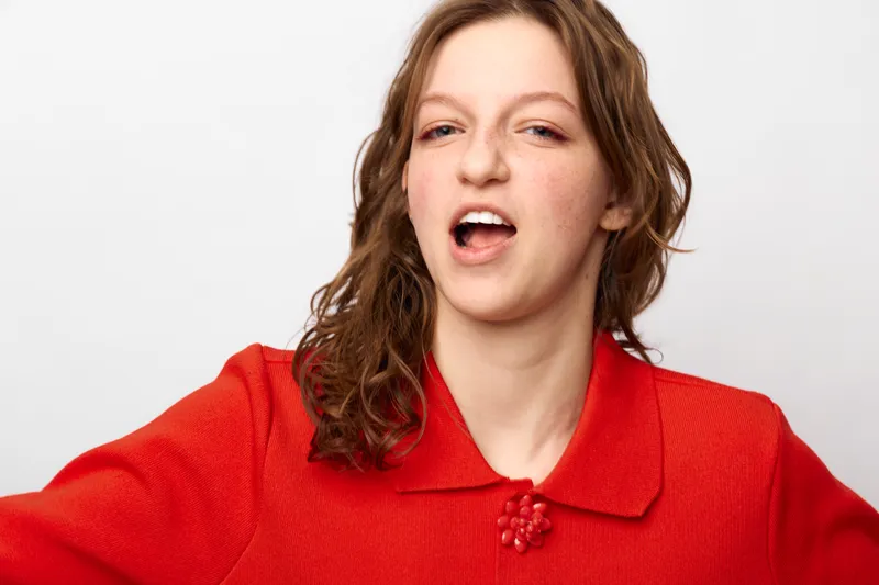 Young woman with long brown hair wearing a red sweater makes a playful face against a white background. Casual portrait with natural expression and relaxed mood in studio lighting.