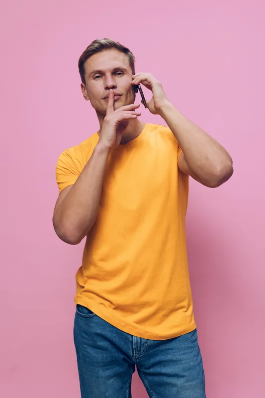 man with smartphone making silence gesture holding phone in orange t-shirt isolated pink background digital detox concept