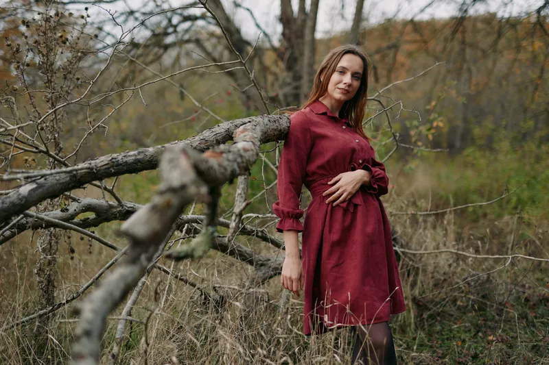 Candid portrait of a woman in a simple red dress leaning on a fallen tree in a grassy meadow, showing genuine natural presence, subtle emotion and real lifestyle mood, quiet confidence.