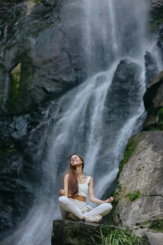 Young woman meditating in lotus pose near a powerful waterfall, wearing white athletic wear, surrounded by natural rocks and greenery, embodying peace and mindfulness.