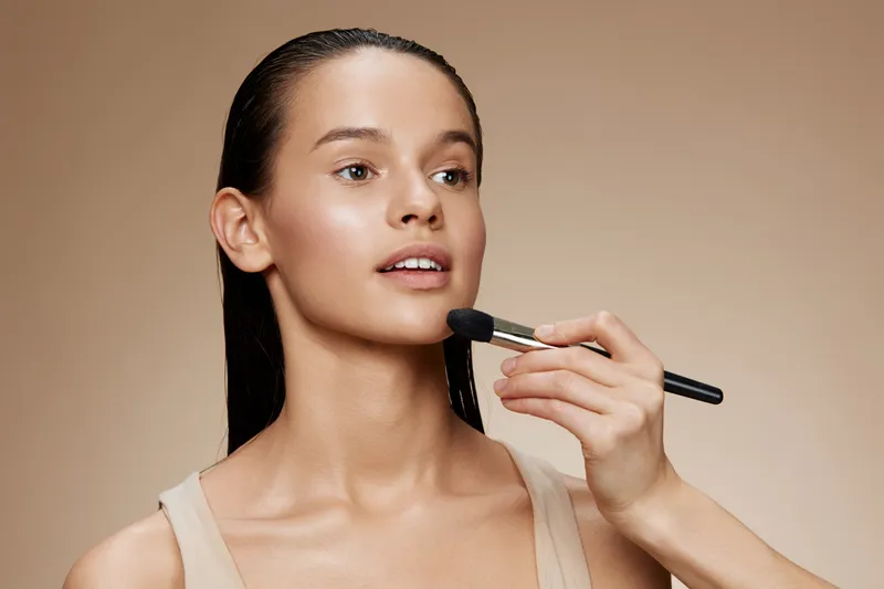 Young woman receiving makeup application with a natural look, showcasing beauty and skincare, neutral background, soft lighting, and warm color palette
