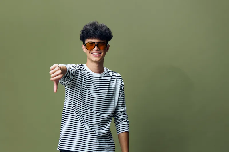 young man wearing sunglasses and striped shirt showing thumbs down gesture with a confident smile, olive green background, casual and playful expression