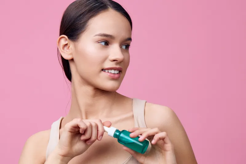 Young Asian woman applying serum on her skin, smiling, with a pink background showcasing skincare routine and healthy beauty concepts