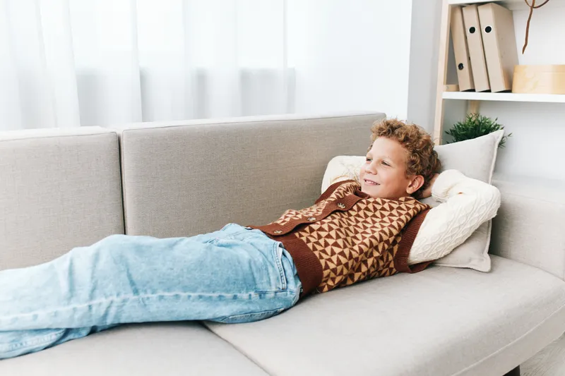 Young boy relaxing on a light sofa, smiling contently as he lounges with cushions, wearing a patterned sweater and blue jeans, in a bright living room.