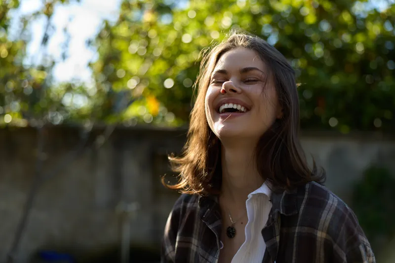 Vibrant woman outdoors, smiling with eyes closed, sunlit moment, casual lifestyle, natural lighting, lush greenery background, relaxed posture, authentic joy, candid portrait vibe