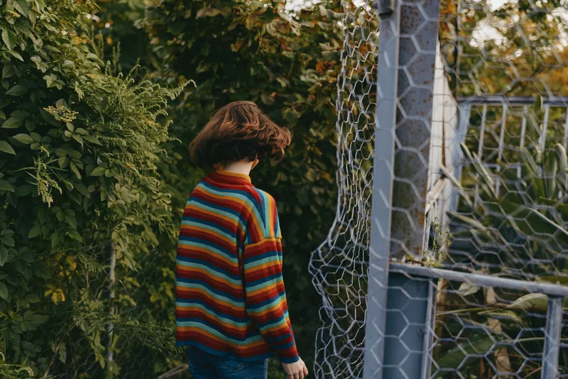 Young woman in a colorful striped sweater walking through a lush garden, back view showing short hair and casual outfit near a wire fence, relaxed contemplative mood.