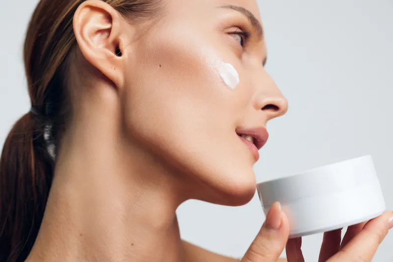 Young woman applies face cream on cheek holds white jar close to skin skincare beauty routine isolated on white background natural healthy skin care concept.