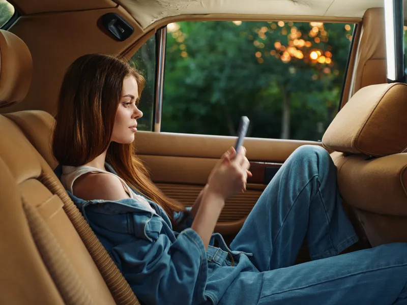 Young woman in denim outfit relaxing in car, using smartphone, with nature background during sunset, representing comfort and modern lifestyle.