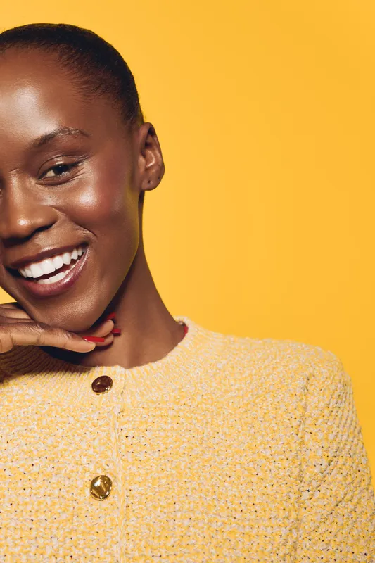 Smiling African American woman with dark skin poses with hand near chin wearing yellow textured jacket. Bright yellow background creates vibrant mood, concept of beauty, confidence, and fashion.