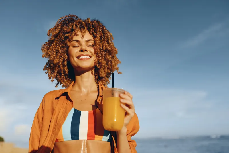 Smiling woman with curly hair on a sunny beach holds a bottle of juice, wearing a striped swimsuit and orange shirt, enjoying a bright summer day by the sea.