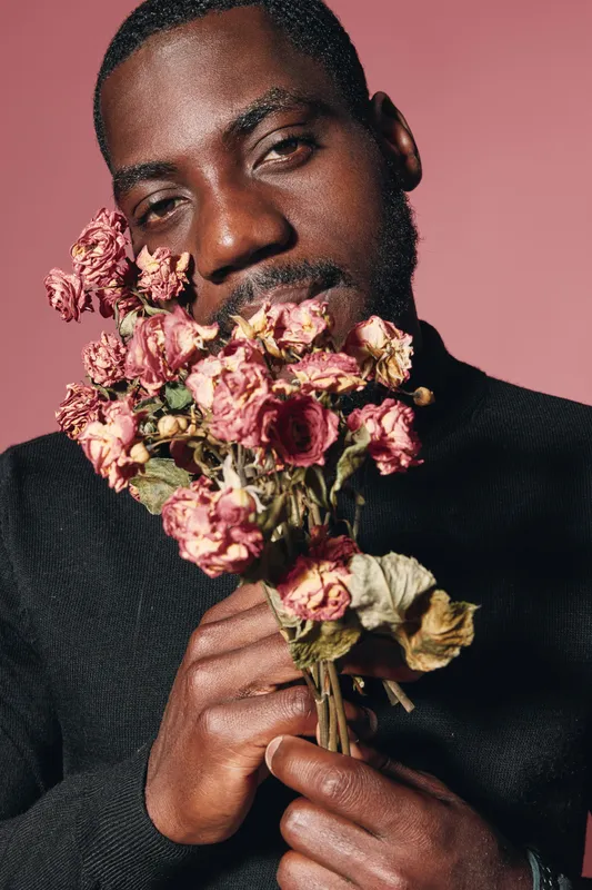 African man holding dried pink flowers close to face in creative conceptual visual style with dark skin and black sweater on pink background.