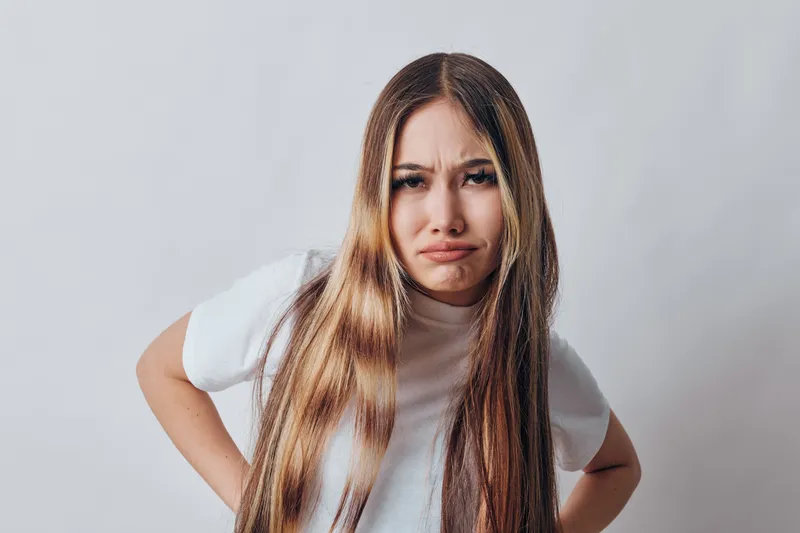 Young woman with long hair frowning with discontent standing with hands on hips isolated on white background. Female expressing disappointment or frustration with unhappy facial expression.