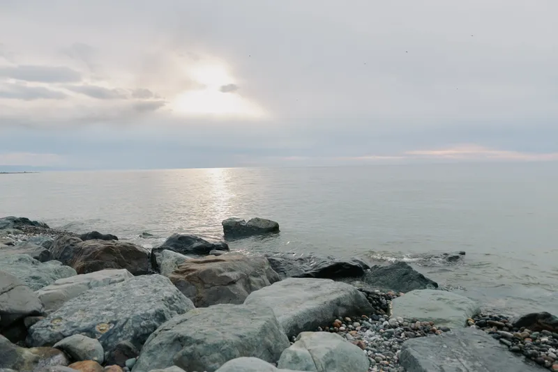 Rocky shore at calm sea under cloudy sky with sun reflection on water surface. Peaceful coastal landscape in soft natural light. Concept of nature, tranquility, and ocean view.