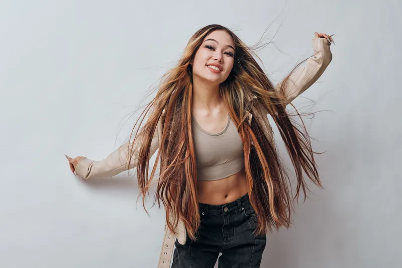 Young woman with long hair and casual clothes smiling and posing with raised arms against plain background for lifestyle and freedom concept.