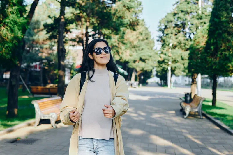Fashion outdoors park scene with sunglasses and coat, casual style and sunny daytime energy, a woman walking along a tree lined path in urban mood and leisure vibe