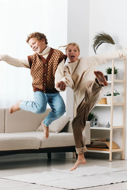 Two playful children jump mid air indoors, smiling and carefree as they wear cozy knitwear and jeans. Bright living room setting conveys energy, joy, and spontaneous fun.
