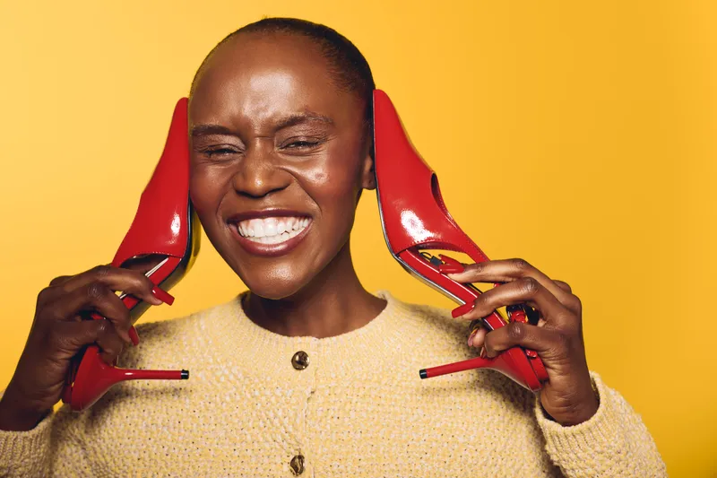 Smiling African woman with dark skin holds red high heels near her ears on yellow background. Concept of fashion, happiness, style, and beauty with vibrant colors and joyful expression.
