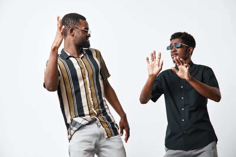 Two black men with dark skin wearing sunglasses dancing and having fun together in casual summer clothes on white background in studio.