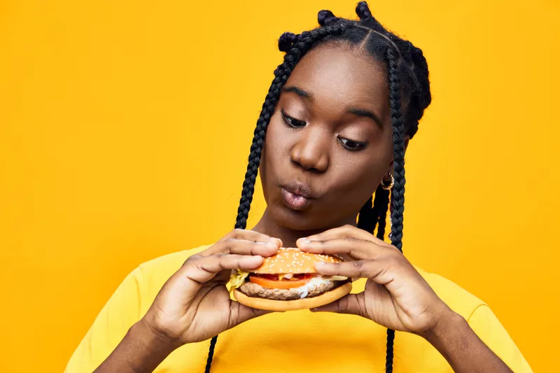 Young Black woman enjoying a delicious hamburger, showcasing excitement and curiosity against a vibrant yellow background, perfect for healthy eating concepts