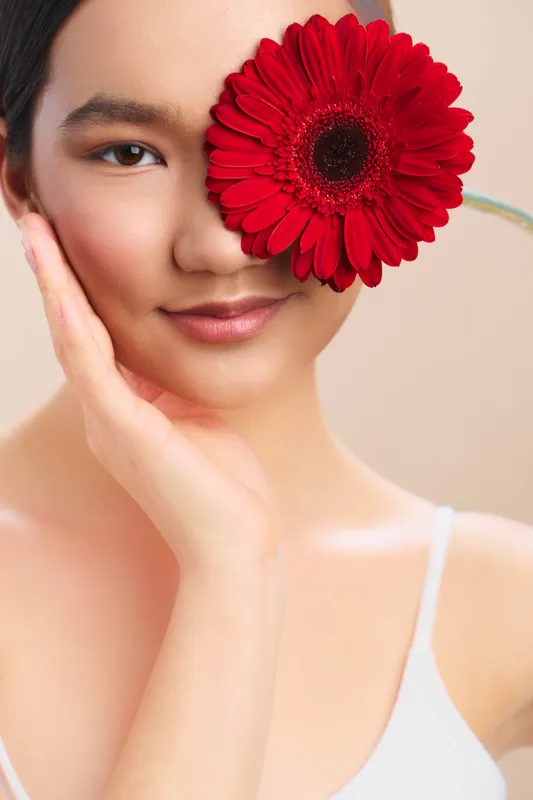 asian woman with a red flower covering one eye, closeup portrait with smooth skin, soft lighting, and a warm smile creating a graceful, artistic mood.