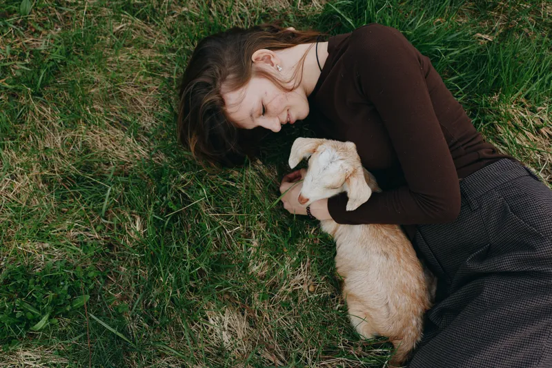 Joyful woman outdoors, lying on grass, cradling a gentle goat kid, warm sunset light, relaxed pose, natural textures, soft breeze, earthy tones, peaceful bond with nature, candid lifestyle moment