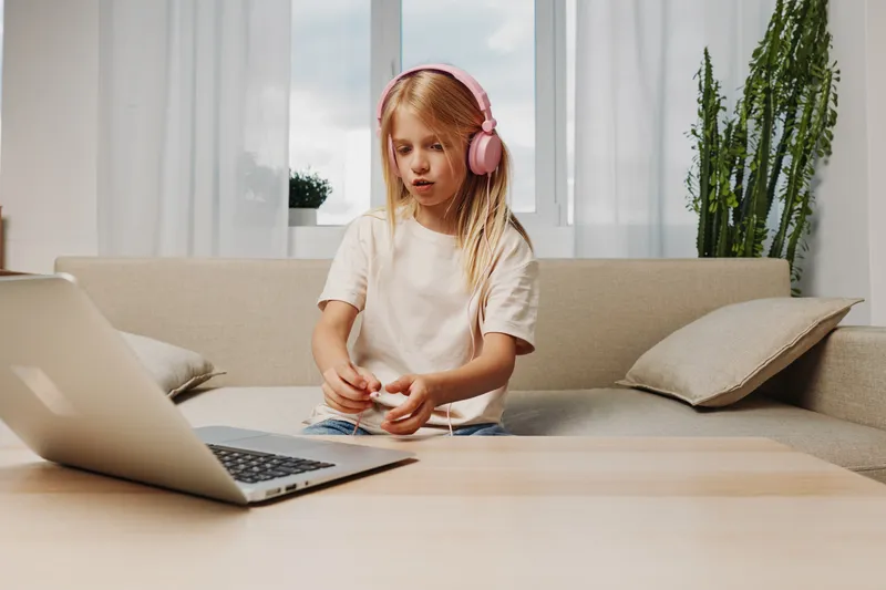 Blonde girl with pink headphones focuses on taking a pill while sitting on couch near open laptop in bright living room. Concept for child health, remote learning, and modern lifestyle.