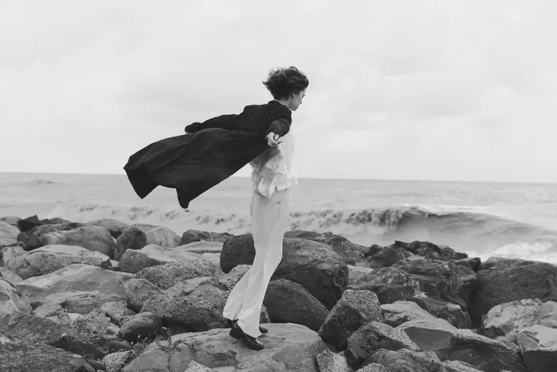 Couple on rugged rocks by the sea, one lifting the other in a flowing cloak, wind catching fabric, serene motion against the horizon.