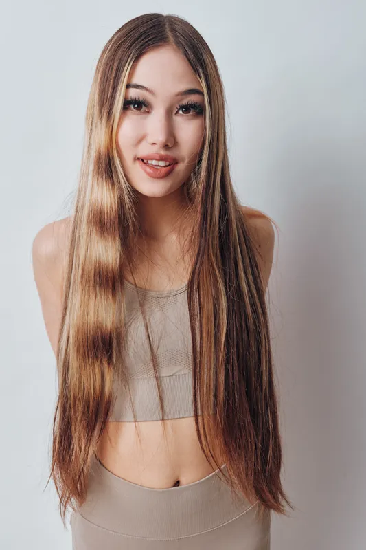 Young woman with long straight hair smiling, wearing beige top and standing against a light background. Female model with natural makeup and casual outfit in studio.