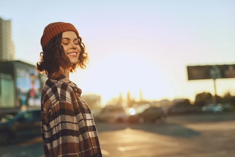 woman smile portrait golden hour urban beanie candid happiness: Authentic candid lifestyle portrait of a smiling young woman in plaid coat at sunset, warm golden glow, mindful living and emotional