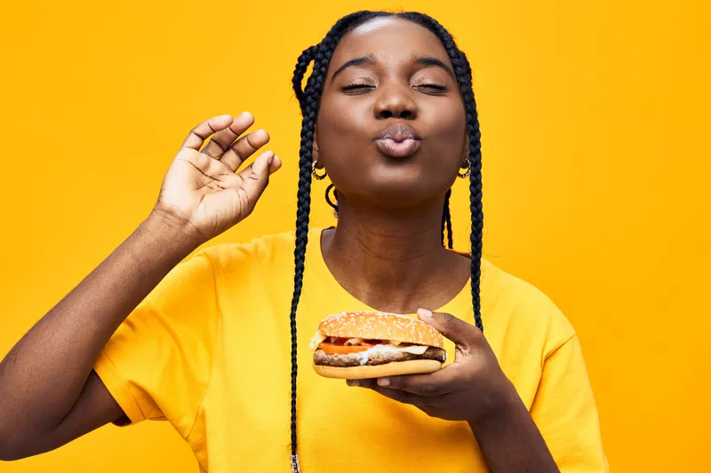 Young Black woman enjoying a delicious burger against a bright yellow background, expressing joy and satisfaction with a playful kiss gesture