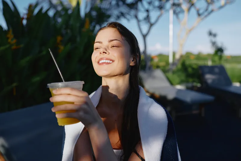 Cheerful woman outdoors, sunlit moment, holding a beverage by poolside, towel draped over shoulders, relaxed posture, vibrant greenery background, soft breeze, natural colors