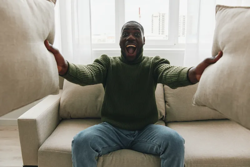 Happy man celebrating with cushions in a cozy living room, wearing a green sweater and jeans, showcasing joy and excitement in a bright, modern space