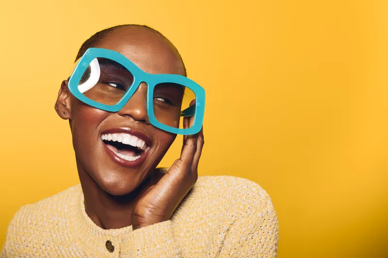Happy African American woman with dark skin wearing large blue glasses and yellow sweater laughs joyfully on yellow background. Concept of fashion, happiness, and positive emotions.