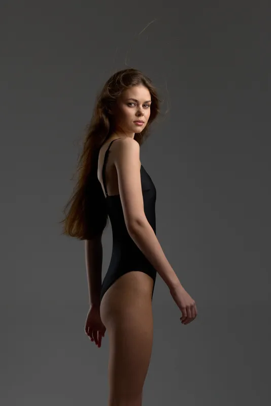 Young woman in black bodysuit posing confidently against gray background with serious expression and long hair.