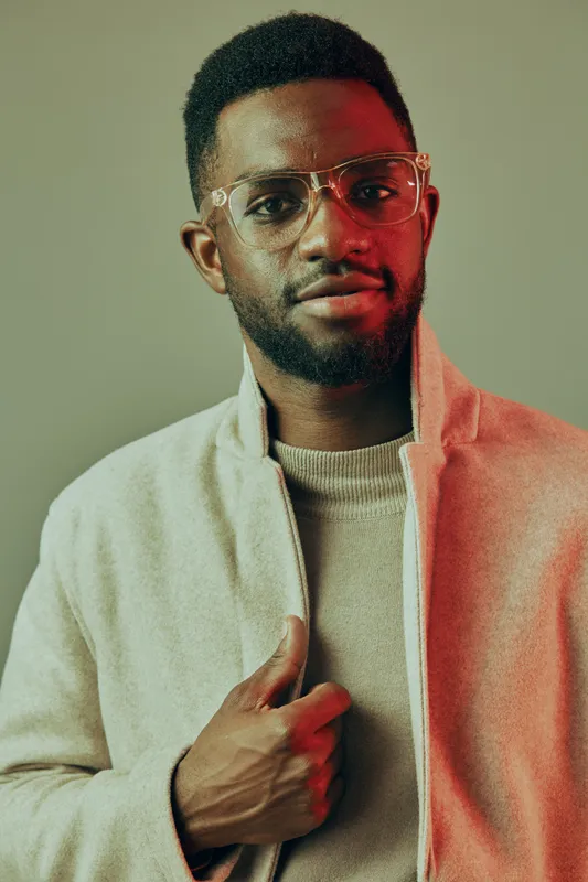 Confident Black man in stylish glasses and modern attire showcases fashion forward look against a minimalistic background, emphasizing self expression and individuality