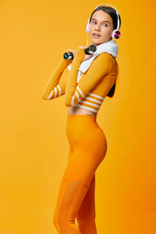 Fit young woman in orange seamless workout set holds dumbbells and towel, wearing headphones, posing against bright studio backdrop for modern sport and fitness lifestyle imagery.